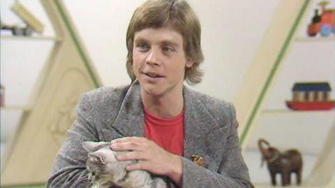 Mark Hamill in the Blue Peter studio holding a cat, wearing a red T-shirt and grey suit jacket.  Set design in background shows an elephant and a boat.
