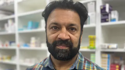 Man wearing a checked shirt with shelves behind containing small packets.