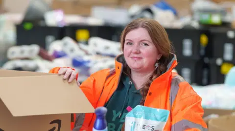 A woman with ginger hair, dressed in a orange hi-vis jacket and a green hoodie, with her hand resting on a cardboard box in a warehouse 