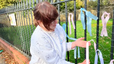 Adele Mann wearing a white/grey jumper covered in teddy bear faces, standing next to a metal fencing with ribbons on it - holding a pink ribbon