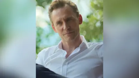 A man in a white shirt with the collar unbuttoned sits with his legs crossed