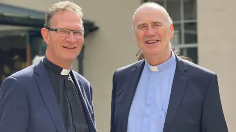 Peter O'Reilly/PA Wire Two men in blue suits wearing dog collars. The man on the left is wearing glasses, with black curly hair. The man on the right has grey hair.