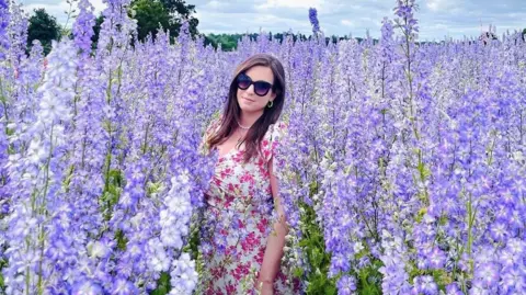 Thedopaminediaries_ A woman wearing a white and pink floral dress middle of a flower field. Dozens of blooming lilac and purple flowers are around her.