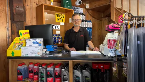 Martin Gillott stands behind his shop till. Around him is car equipment: number plates, windscreen wash and car air fresheners
