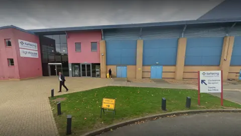 Google Pink entrance to theatre building with blue and brown exterior walls. There is a small grass patch in the foreground. A man in a dark suit is walking past.