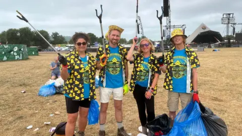 Angela Saxbee, Alastair Cullen, Sarah Parker and Gavin Eddy. Angela has dark curly hair tied up in a ponytail, Alastair has short fair hair and facial hair, Sarah has short blonde hair and Gavin has short blonde hair. Alastair and Gavin are wearing yellow hats, while Angela and Sarah are wearing sunglasses. All of them are wearing blue volunteer t-shirts and black shirts with yellow ducks on them. They are all holding litter picking sticks and bags of rubbish.