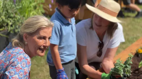 HRH the Duchess of Edinburgh smiles on the left side of the left of the picture. She is wearing a blue dress with red detail. In the middle there is a young pupil with black hair wearing a blue polo shirt and gardening gloves. On the right a woman is wearing a straw hat with a white t-shirt and sunglasses in the collar. 