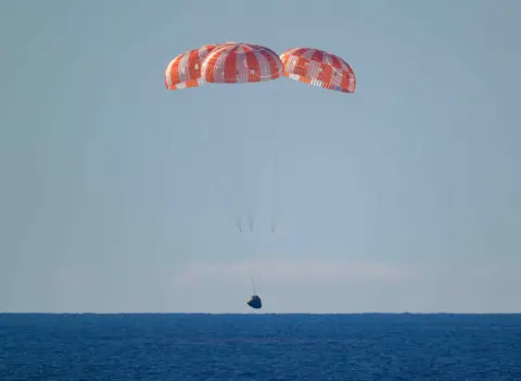 NASA The landing craft with it's orange parachutes lands on a dark blue sea.