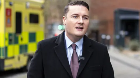 Wes Streeting in shirt tie and jacket standing in front of an ambulance.