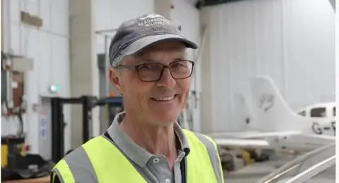 BBC A man in a cap and hi-vis jacket. He has glasses and is smiling at the camera. He is standing inside a warehouse building where aeroplanes are kept.
