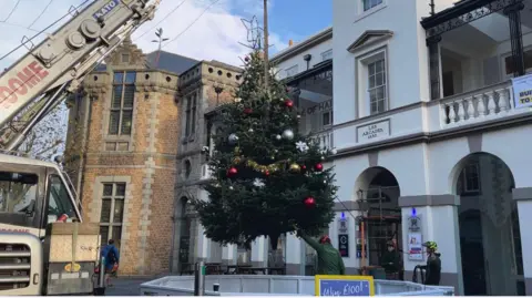 A Christmas tree decorated in red, silver and gold baubles, gold tinsel and white snowflake decorations. It has a star on the top. The tree is being lifted by crane above a white barrier, guided by a man in an orange helmet and green fleece. 