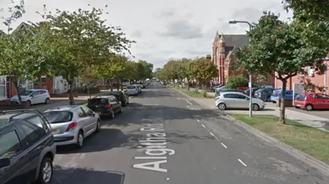 Google A long, straight and wide road is flanked by trees and houses. Cars are parked on the side of the road.