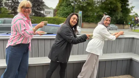 Aisha Iqbal/BBC Three women lined up and posing in running mode while holding teacups and saucers. They are all middle aged. Two look Asian, the other is white. One of the Asian ladies wears a headscarf.