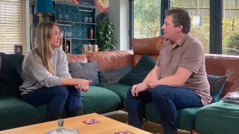 Mousumi Bakshi/BBC Georgina Bayliss with long blond hair wearing a grey sweater and blue trousers sitting on a green sofa with her husband, Scott, who has medium-length dark hair and is wearing a brown T-shirt and blue trousers. There is a coffee table in front of them and wooden shelves behind.