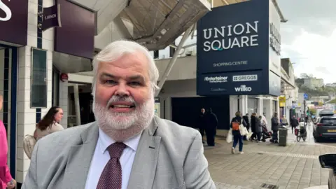 David Thomas, Conservative leader of Torbay Council, wearing a gray jacket and burgundy patterned tie, standing in front of the Union Square sign