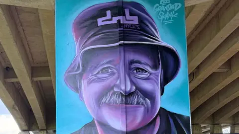 Nathan, a man, standing in front of an artwork on a pillar of a man wearing hat.