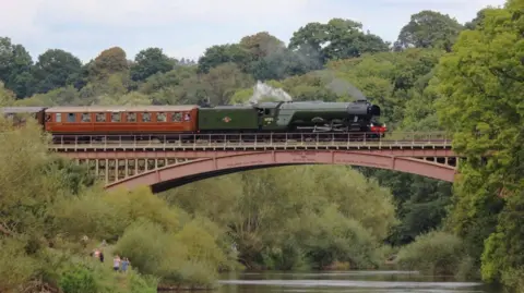 Severn Valley Railway/Keith Wilkinson The largely green-coloured locomotive is on the bridge in the middle of the photo, with one of the carriages fully visible (from left to right) on the left of the photo. Trees are in the background.