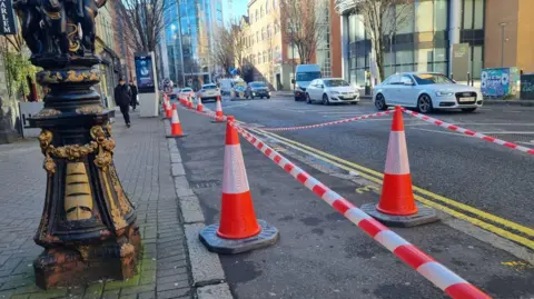 BBC Rows of fluorescent orange cones with tape strung between them on a footpath along Bedford Street in Belfast