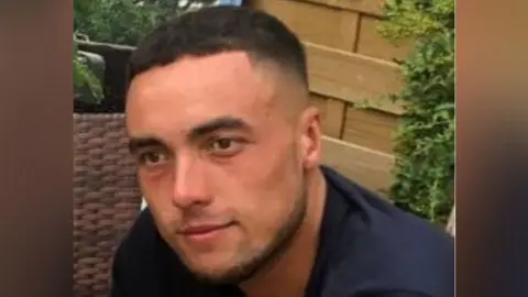 Kieran Shepherd sitting outside wearing a navy shirt. He has black hair and has a faint smile in the photo.