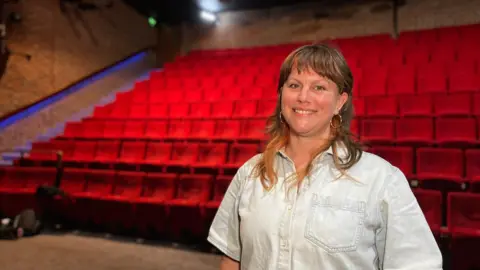 Esther smiles at the camera as she wears a short sleeved, light blue denim shirt. She has short brown hair which comes to her shoulders. Behind her are red theatre seats.