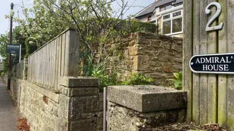 A wall has a wooden fence on to of it and at the end there is a for sale sign under some young trees with blossom on them. A second wall block the view of a house but you can just see an upstiars window and the roof and there is a sign which says Admiral House. 