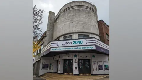 Luton Borough Council An empty Art Deco cinema in Luton. It shows a building, with light curved brick, with a board with Luton 2040 written on it.  There are windows above and two main doors to the building. There are signs around the front door.