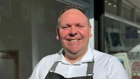 An image of the owner of Hamilton's Tea House, Steve Hamilton. He is looking at the camera and smiling. He is wearing a white top covered by an apron.