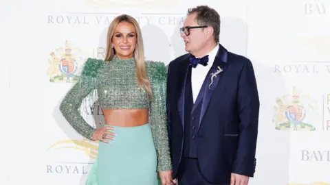 PA Media Amanda Holden poses with a hand on her hip on the red carpet. She's wearing a green sequinned crop top and green skirt, and is standing next to Alan Carr who is wearing a shiny navy suit and is turned to look at her.