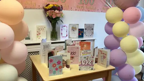 GUY CAMPBELL/BBC  100th birthday cards can be seen on a wooden table with yellow, purple and pink balloons either side. 