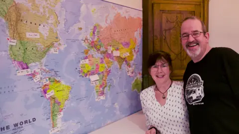 BBC A woman with brown hair and glasses wearing a white top with blue markings next to a man in a black t-shirt with a camper van logo. They are stood next to a world map
