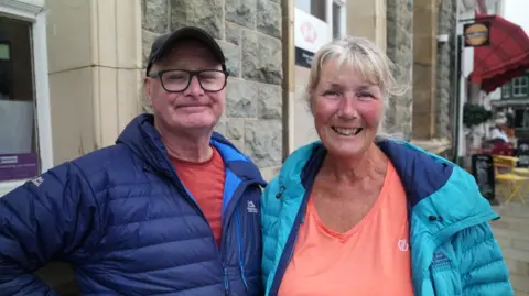 The husband and wife are in blue coats smiling standing in front of a cafe. Chris has white hair and a fringe, and wears an orange t-shirt. Shaun wears glasses, a cap, and a red t-shirt. 
