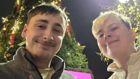 Parker Billy and Parker pose for a selfie at night in front of a large Christmas tree decorated with lights and ornaments. One person is wearing a grey fleece jacket and the other a light-coloured zip-up jacket. Bright streetlights illuminate the scene, and festive lights can be seen on the tree and surrounding decorations in the background. A partially visible pink sign appears behind them near the base of the tree.