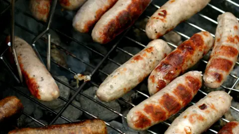 BBC Sausages cooking on a barbecue with one being held by a tong