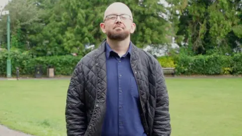 A bald and beared bespectacled man in his thirties, wearing a blue shirt and trousers and a dark jacket, stands in a park with his hands in his pockets.