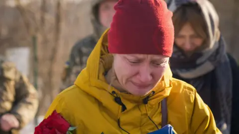 Matthew Goddard/BBC Natalia weeps by the grave of her husband in a snow-covered cemetery in Kyiv. She's dressed in a yellow jacket and a red beanie hat, and is holding a red rose. 