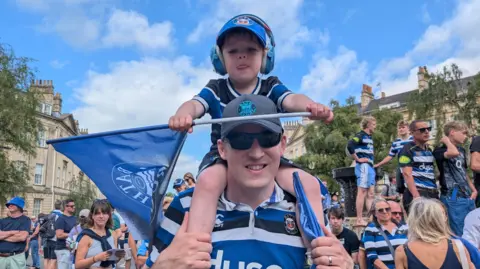 A boy with ear defenders perched on a man's shoulders. They both have Bath shirts, hats and flags