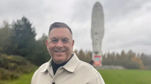 Gary Conley, who wears a white jacket, smiles while standing in front of The Dream sculpture on a green mound. It is made of white stone, which shows a head of a girl with a ponytail. Trees surround it in the background.