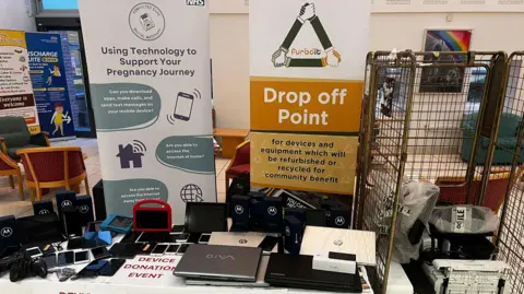 A table containing a number of electronic devices - mostly laptops and mobile phones. It is backed by a sign saying "Drop off Point for devices and equipment which will be refurbished or recycled for community benefit" and another stating "Using Technology to Support your Pregnancy Journey".