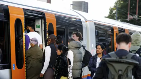 Passengers board a crowded London Overground train, with people queuing at the open doors of an orange-and-white carriage.