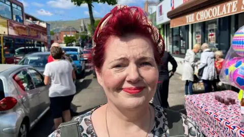 Imelda is looking at the camera and smiling. She is wearing red lipstick and has red short hair. She is wearing a head scarf.
