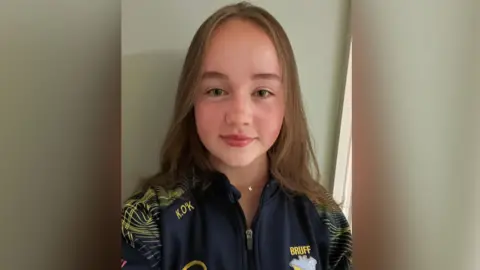 Chris Culhane A young girl with shoulder length brown hair is smiling into the camera. She is wearing a zip up navy and yellow tracksuit top.