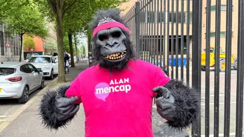 A person stands on a sidewalk wearing a full gorilla costume with a black mask, furry arms, and a bright pink headband topped with a small tiara. The person points both hands toward a bright pink T‑shirt that reads “ALAN mencap” in white text. Parked cars, trees, metal railings, and brick buildings line the street in the background.

