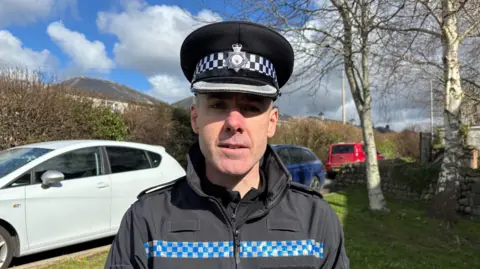 A male police superintendent dressed in police uniform looks at the camera in front of a car park