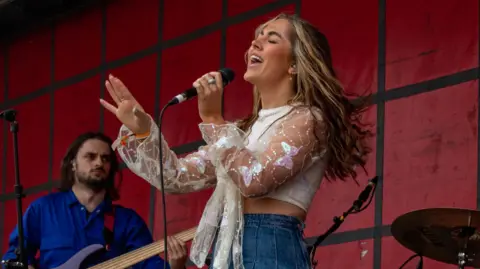 MACY MACY singing on stage while holding a microphone. Her bassist, a man, stands behind her.