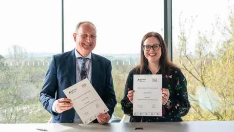 UCS A man in a blue suit and a woman in a black patterned dress, both smiling, holding up signed headed paper reading memorandum of understanding, standing in front of large glass windows with trees outside