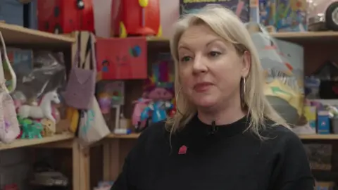 Sophie Livingstone seated indoors in front of shelves filled with toys, books and colourful items, wearing a dark top with a small badge, in a close interview shot.