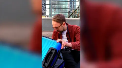 Harry Kind crouches down on the floor as he opens a blue suitcase.