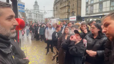 A security guard looks out at a large crowd of people in a long queue in a town centre. Some have hoods up, others carry umbrellas.