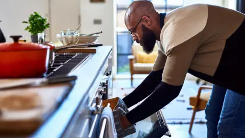 Man with his hands in oven gloves bends down to get something out of the oven, with the work surface and hob in front of him in the kitchen