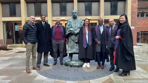 A posed group shot featuring students who took part in the specialist kidney training, together with consultants who created the court. They are five men and two women lining up either side of a metal statue outside Newcastle University.
Four of them, two men and two women, wear a graduation gown with a blue and red and blue stole. The other three men are dressed in formal wear.